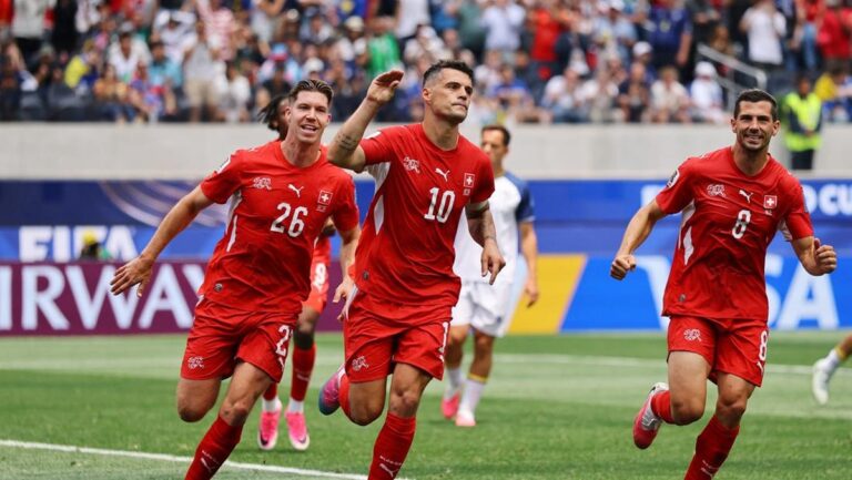 Three Swiss national team players in red jerseys celebrate a goal on a green soccer field as fans cheer in the stands.
