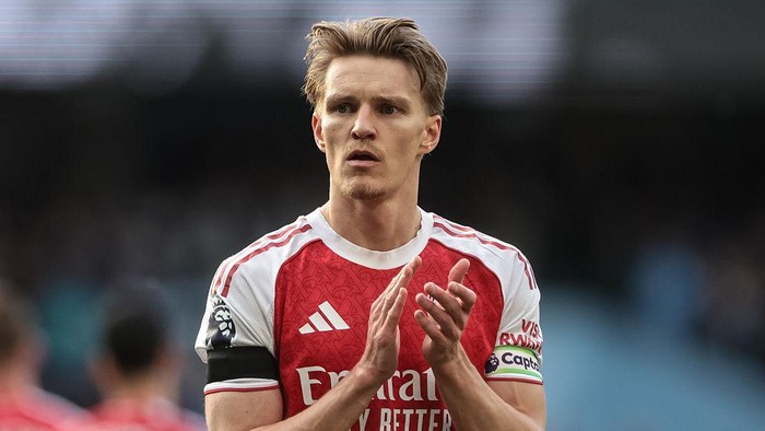 Soccer player in Arsenal jersey clapping on the field, stadium in the background, focused expression.
