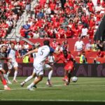 Soccer match scene: red team attacking near the goal defended by a yellow-clad goalkeeper, white team players contest the ball, packed red-clad crowd in stands