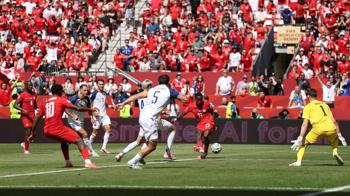 Soccer match scene: red team attacking near the goal defended by a yellow-clad goalkeeper, white team players contest the ball, packed red-clad crowd in stands