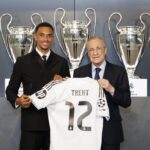 Young footballer and an older official hold up a white jersey that says TRENT 12, in front of a row of large trophy cups.