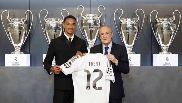 Young footballer and an older official hold up a white jersey that says TRENT 12, in front of a row of large trophy cups.