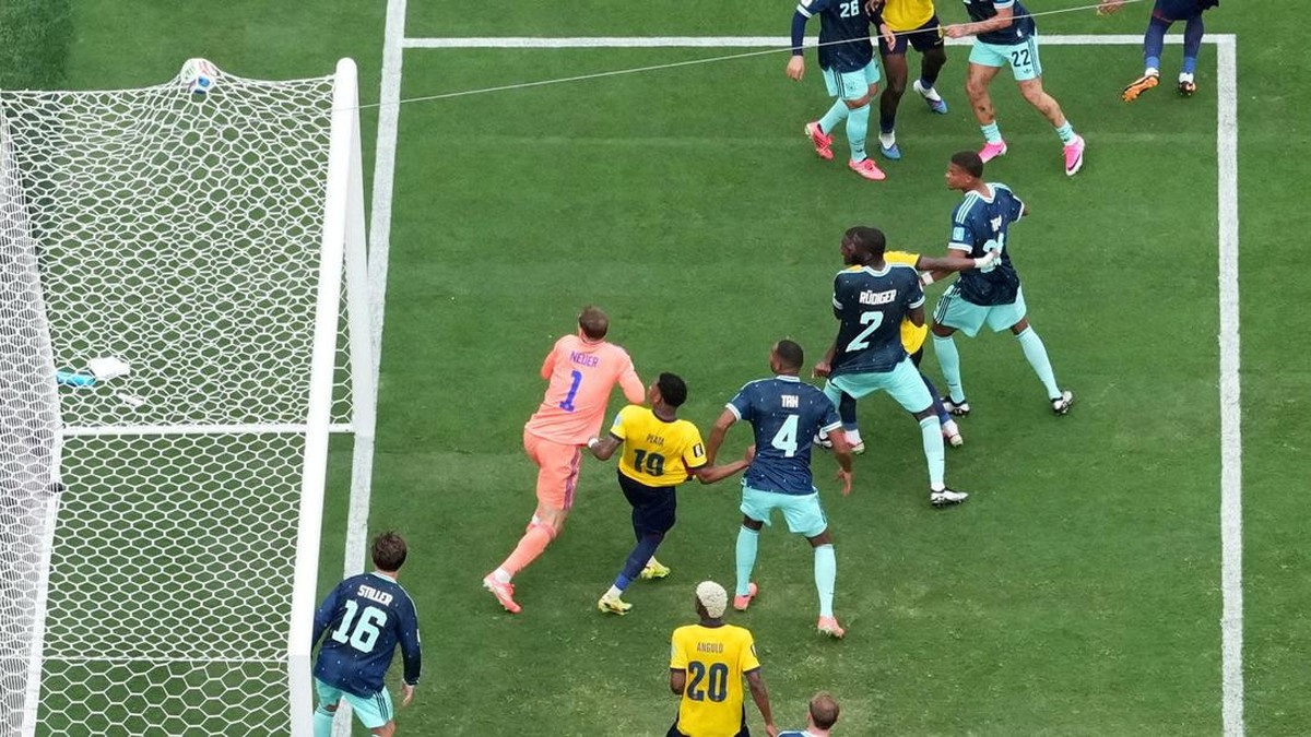 Soccer scene: goalkeeper in pink dives near the goal as players crowd the box and attempt a shot or clearance on a green pitch.