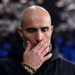 Man with a shaved head covering his mouth with one hand, wearing a dark jacket; appears thoughtful and focused.