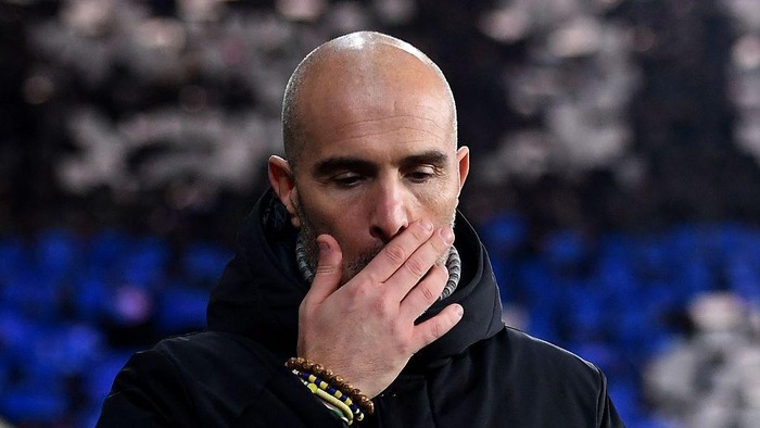 Man with a shaved head covering his mouth with one hand, wearing a dark jacket; appears thoughtful and focused.