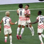 Soccer players in white and red striped jerseys celebrate on the field, jumping together after a goal.