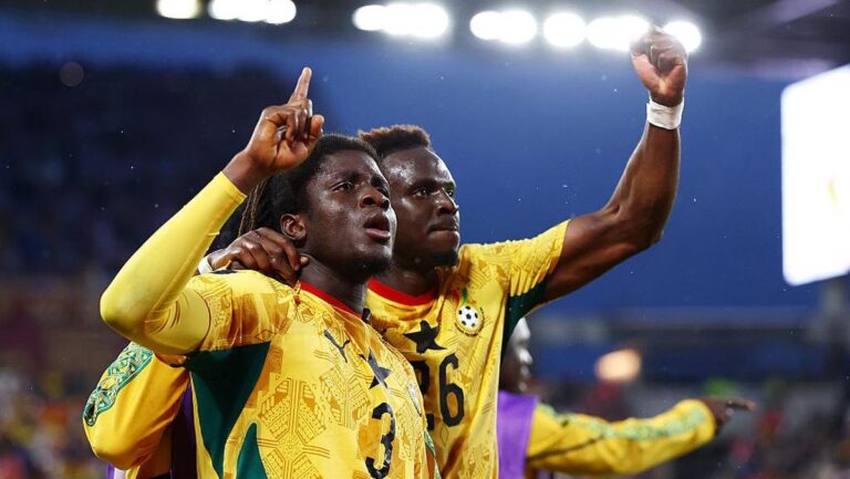 Two football players in yellow jerseys celebrate a goal, one pointing a finger upward while the other wraps an arm around him.