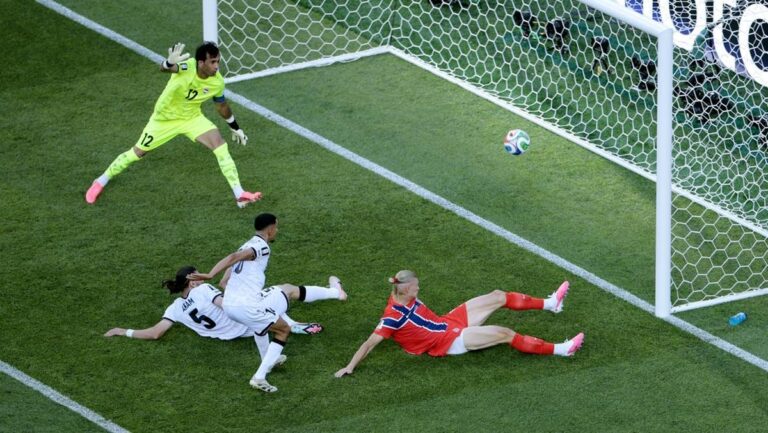 Soccer goalkeeper in a neon kit dives to intercept a ball near the goal as two white-shirt defenders fall to the ground nearby.