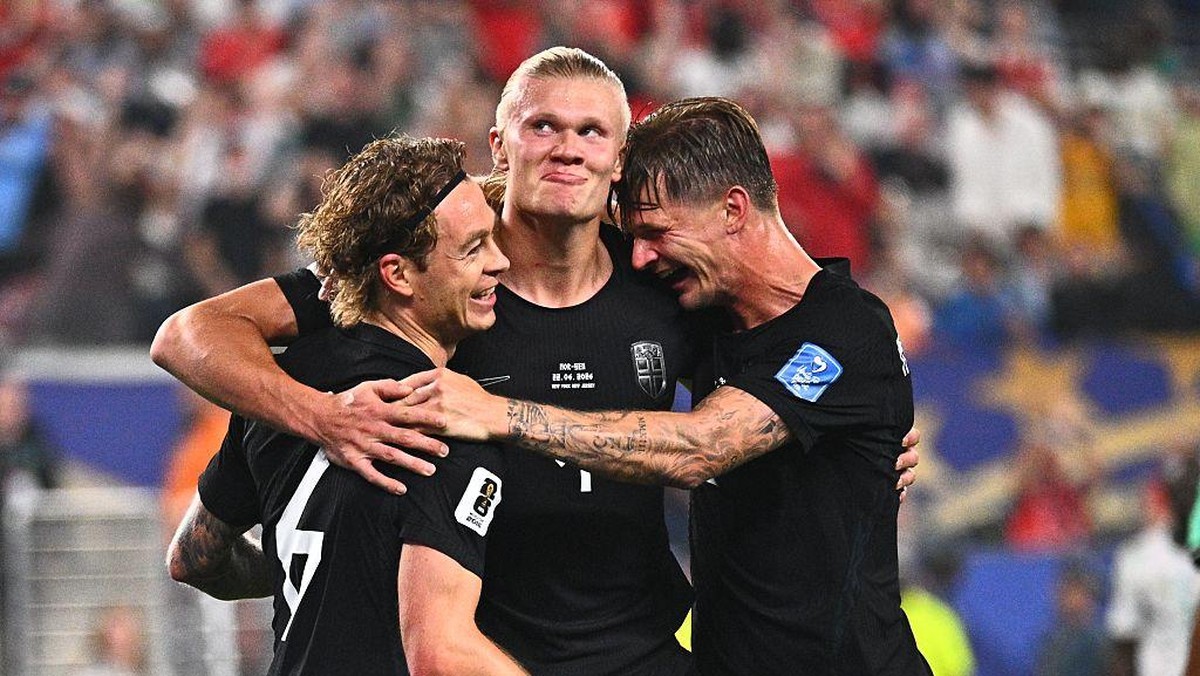 Three male soccer players in black jerseys celebrate with a group hug on the field, stadium blurred in the background.