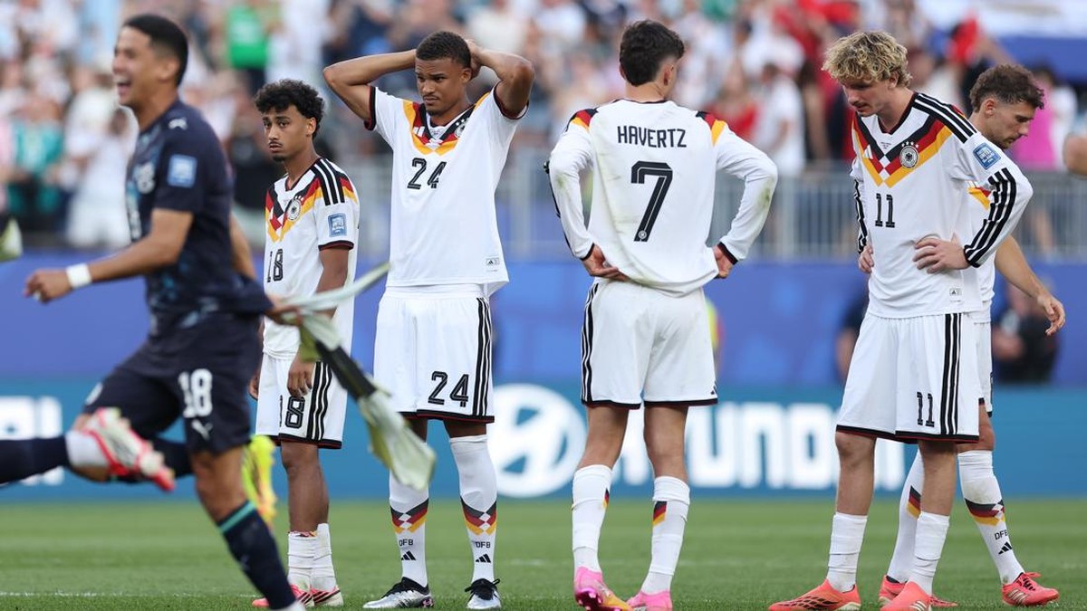 German national team players in white kits with black trim stand on the pitch, Havertz with number 7 visible on his back, arms on hips.