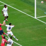 Soccer match moment: a striker in red shoots as teammates and defenders react near the goal with the goalkeeper diving to save nearby.