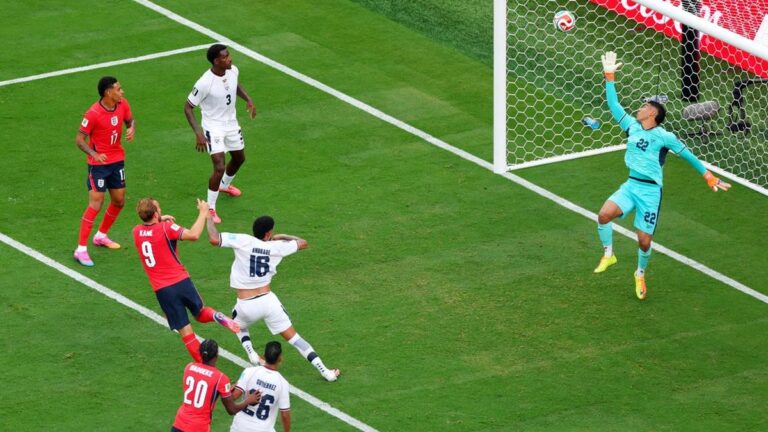 Soccer match moment: a striker in red shoots as teammates and defenders react near the goal with the goalkeeper diving to save nearby.