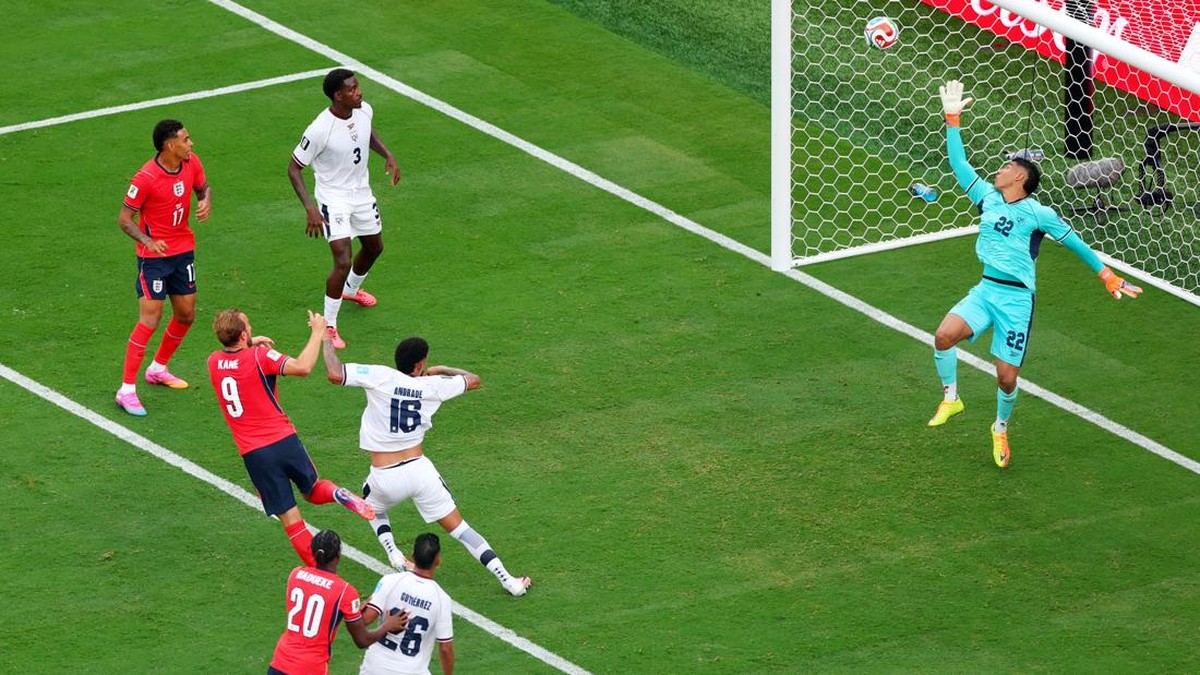 Soccer match moment: a striker in red shoots as teammates and defenders react near the goal with the goalkeeper diving to save nearby.