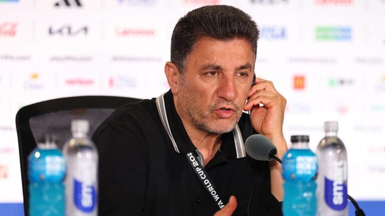Man at a press conference speaking into a microphone, wearing a black polo and lanyard, with water bottles on the table behind him