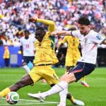 England player in white challenges a Ghanaian in yellow for the ball during a crowded match on the pitch with a packed stadium in the background.