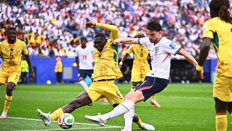 England player in white challenges a Ghanaian in yellow for the ball during a crowded match on the pitch with a packed stadium in the background.