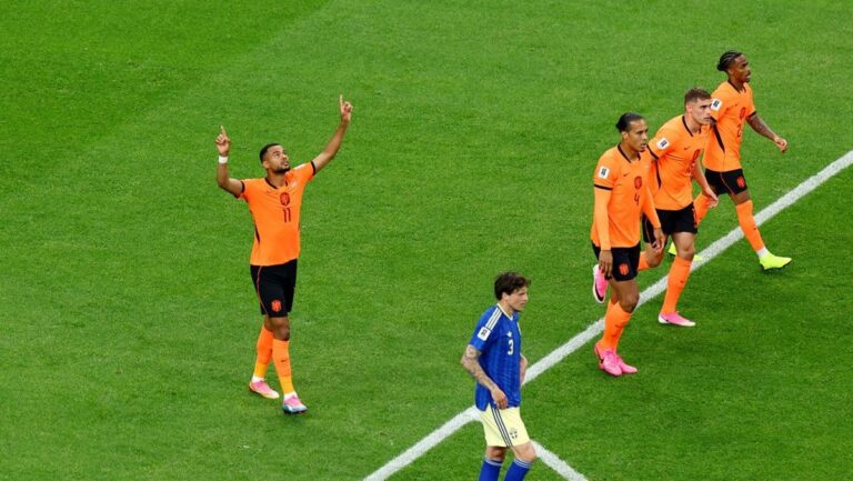 Dutch national team players in orange celebrate on a green soccer pitch after a goal, while a blue opponent looks on nearby.