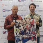 Two men pose holding a colorful sports poster with the caption 'From Jakarta to the dream stage in Japan!!' in front of a sponsor logos backdrop.