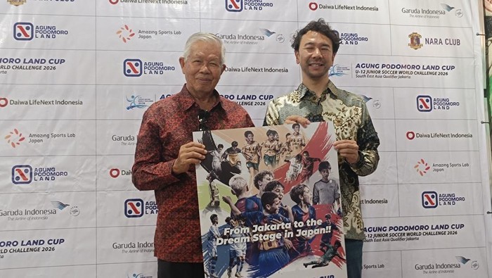 Two men pose holding a colorful sports poster with the caption 'From Jakarta to the dream stage in Japan!!' in front of a sponsor logos backdrop.