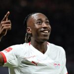 Soccer player in a white jersey celebrates on the field, pointing upward with a smile on his face.