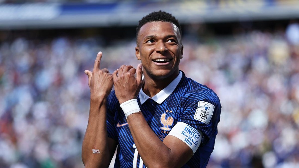 Soccer player in a blue France jersey celebrates on the field, pointing upward with both hands.