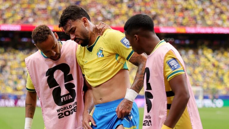 Three Brazilian soccer players on the pitch during a 2018 World Cup match, two wearing pink bibs over yellow jerseys as they support a teammate.