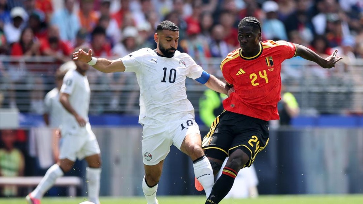Two soccer players contest the ball in a dynamic clash during a match: a white‑jersey player (#10) challenges a red‑jersey defender (#24) in a crowded stadium.
