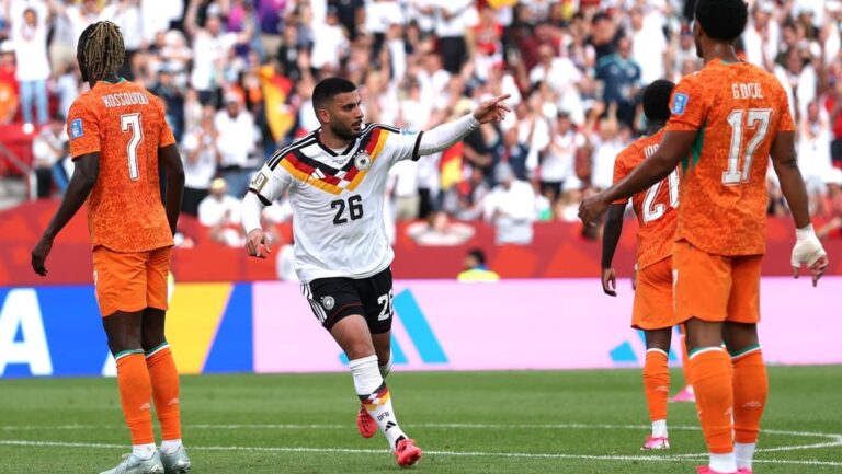 German player (number 26) in white kit points while running on a soccer field, surrounded by orange-clad opponents and a cheering crowd in the stands.