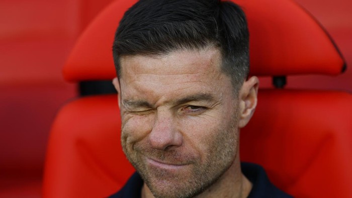 Man with one eye closed in a playful wink, seated against bright red stadium seats in the background.