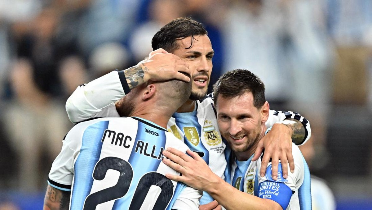 Three Argentina players hug in celebration on the pitch, one wearing a jersey with 'MAC ALLI..' and the number 20 visible on the back, smiling and embracing.