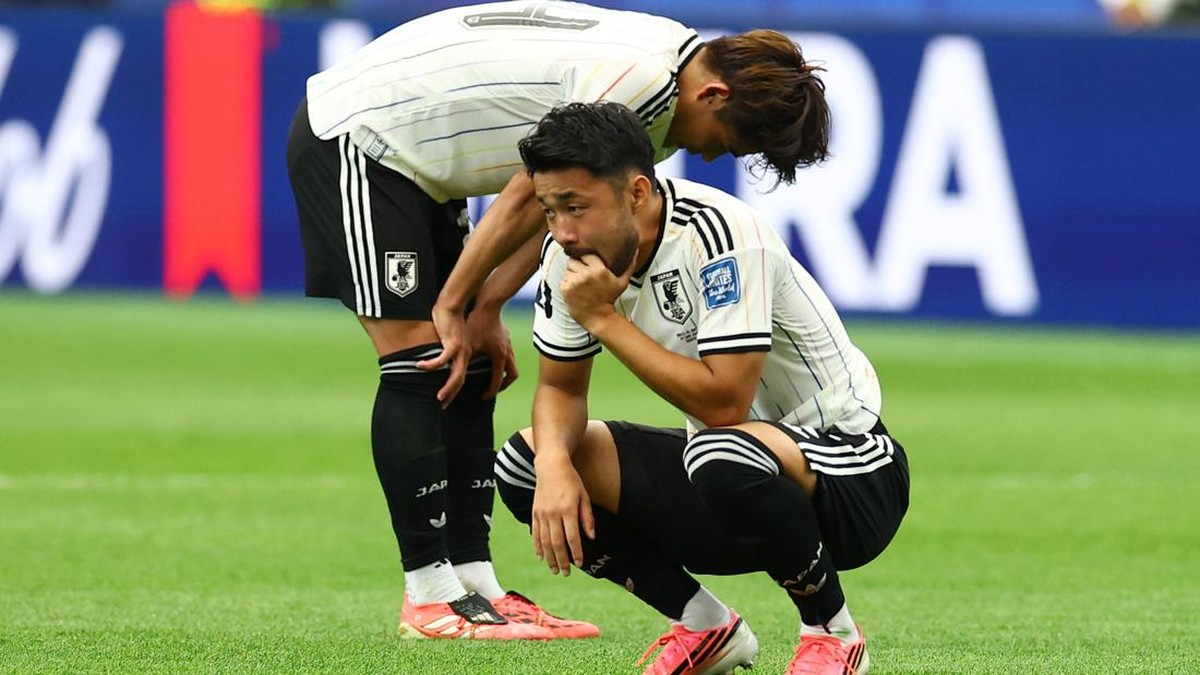 Two soccer players in white jerseys crouch on a green pitch, heads bowed in a moment of reflection after a game.