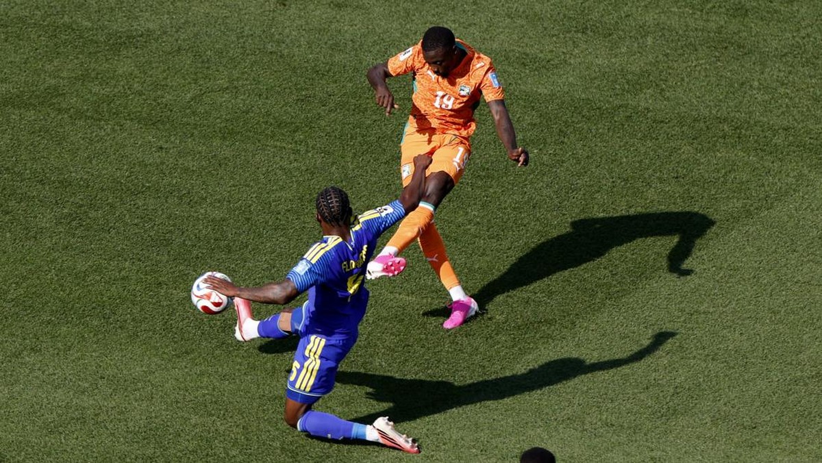 Two soccer players in orange and blue uniforms contest the ball on a green pitch.