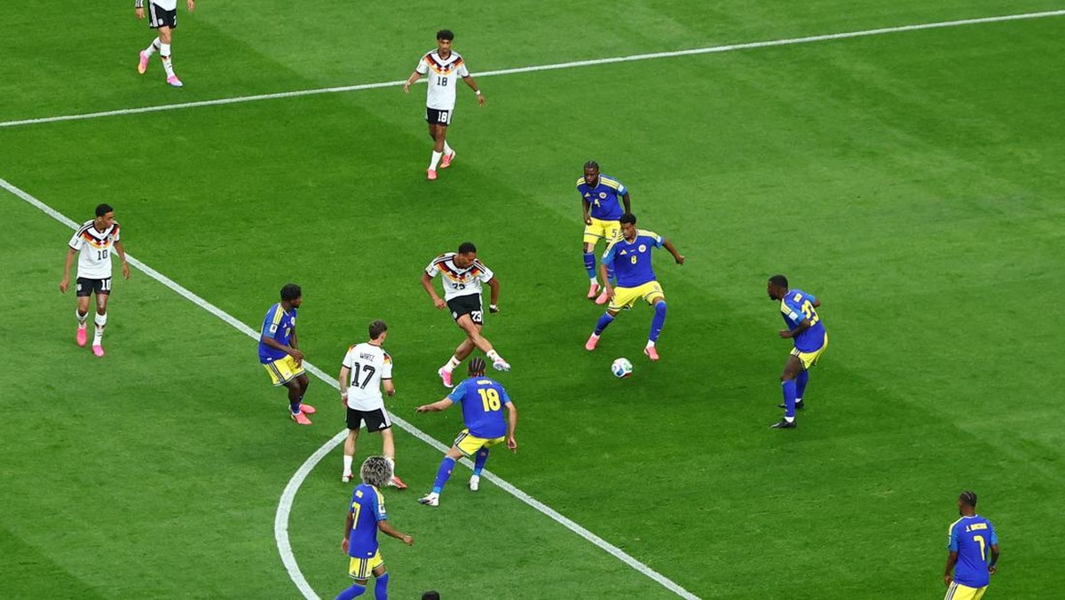 Soccer players from two teams contest the ball in a lively midfield duel on a green pitch.