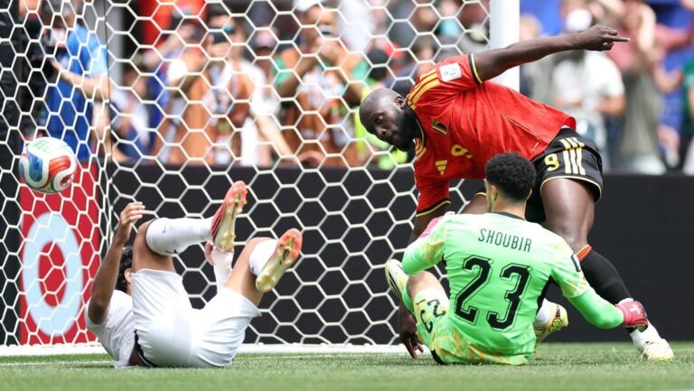 Goalkeeper in green blocks a shot at the goal as a red-clad attacker leans forward near the net with a defender on the ground nearby.