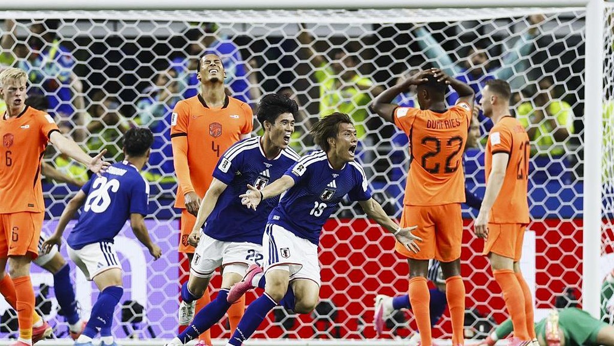 Soccer players in blue and orange jerseys celebrate near the goal after a score, with the net and crowd in the background.