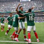 Mexico national team players celebrate a goal, embracing each other on the pitch as fans cheer in the stadium orderly stands behind them.