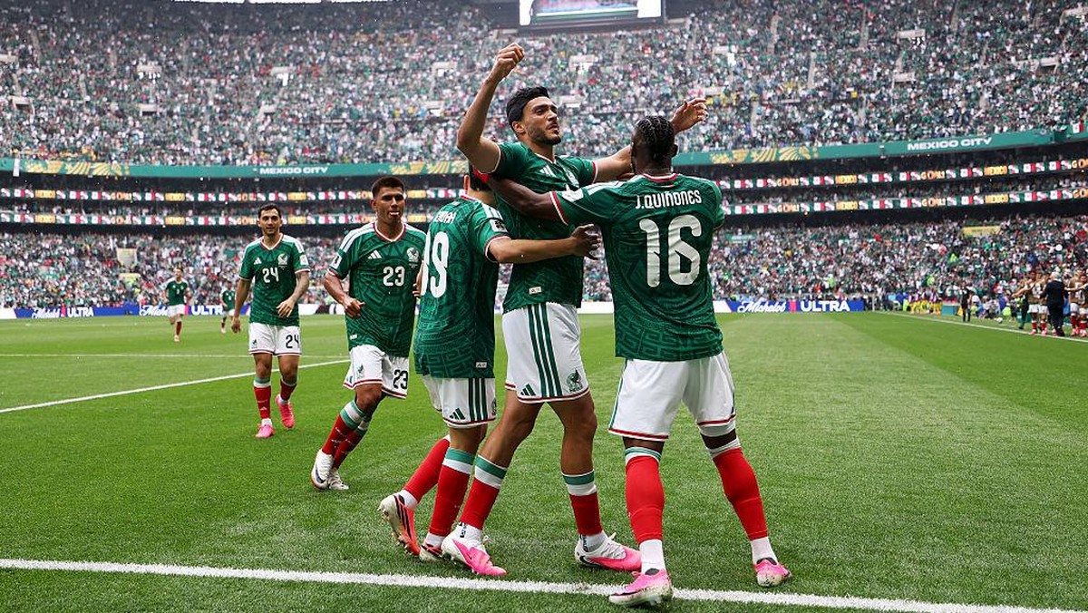 Mexico national team players celebrate a goal, embracing each other on the pitch as fans cheer in the stadium orderly stands behind them.
