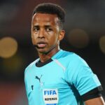 Football referee wearing a turquoise jersey with a FIFA badge and headset mic on a dim stadium backdrop.