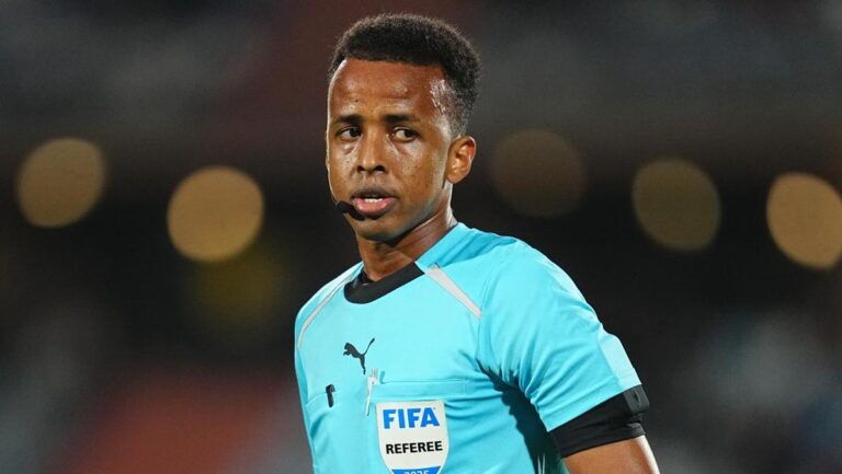 Football referee wearing a turquoise jersey with a FIFA badge and headset mic on a dim stadium backdrop.