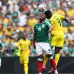 Two soccer players leap to head the ball during a match, with a packed stadium in the background (green jersey vs. yellow jersey).