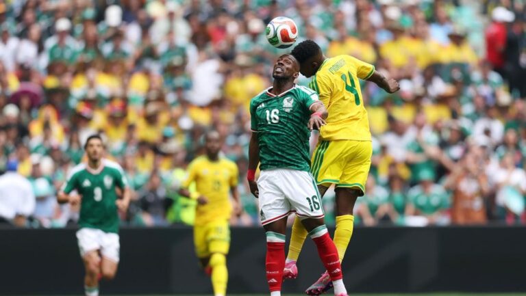 Two soccer players leap to head the ball during a match, with a packed stadium in the background (green jersey vs. yellow jersey).