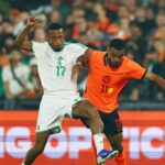 Two soccer players in white and orange kits contest the ball on a crowded field during a match.