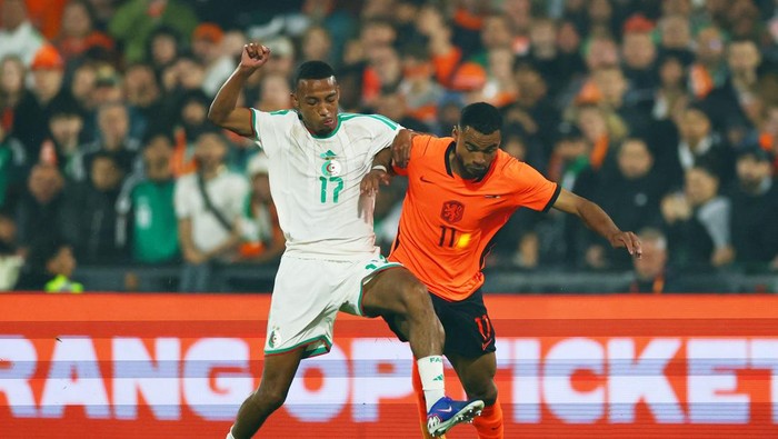 Two soccer players in white and orange kits contest the ball on a crowded field during a match.