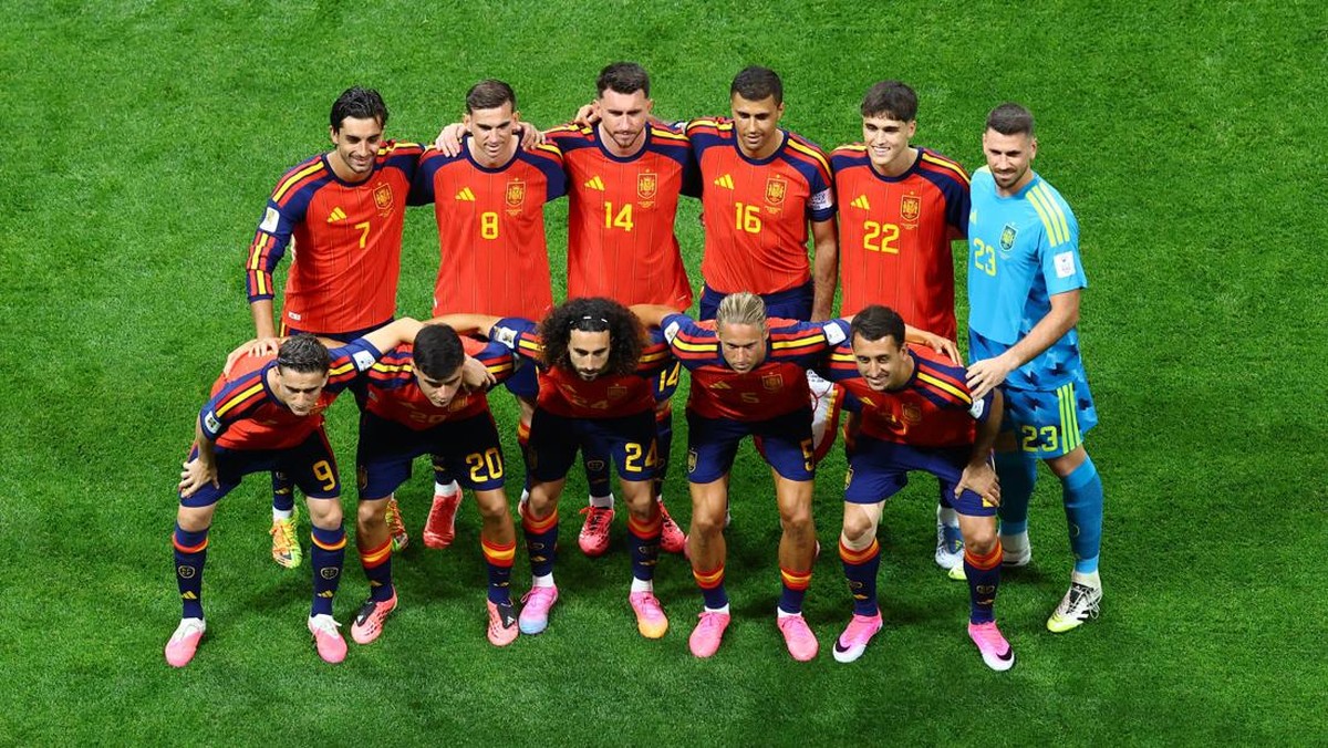 Spanish national soccer team posing in a line on a green field before a match, arms around teammates, goalkeeper in blue at right.