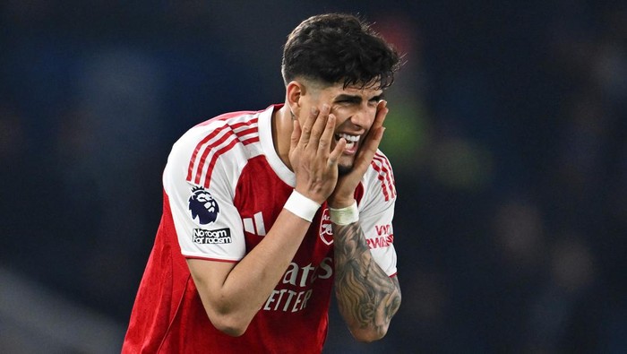 Soccer player in a red Arsenal jersey covers his face with both hands on the field, expressing frustration.