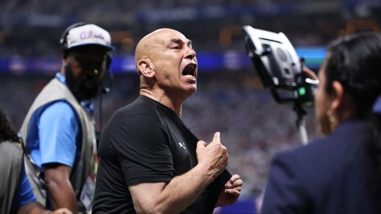 Man in a black athletic shirt shouting passionately on the sidelines of a sports event, fist clenched, stadium crowd in the background.