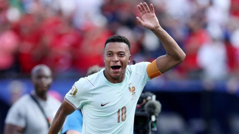 French national team player in light blue jersey with number 10 celebrates on the field, raising his right hand as fans cheer in the background
