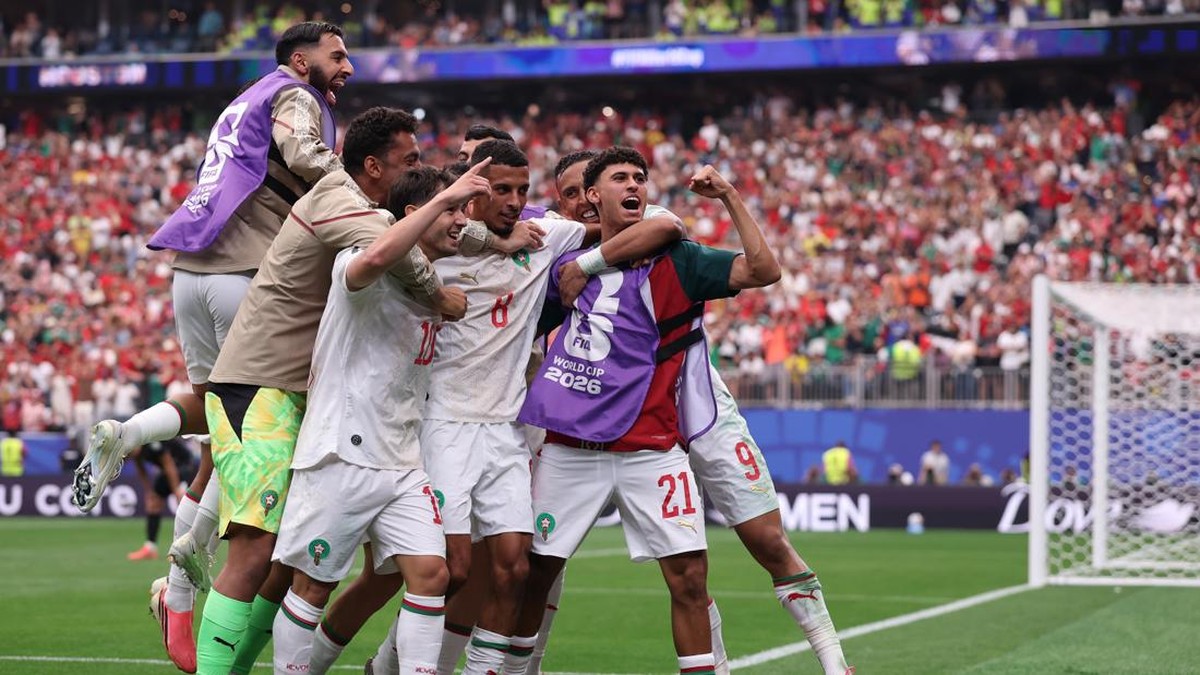 Group of soccer players celebrate a goal on the pitch, wearing white uniforms with red accents and purple World Cup 2026 bibs, as fans cheer in the stands behind them.