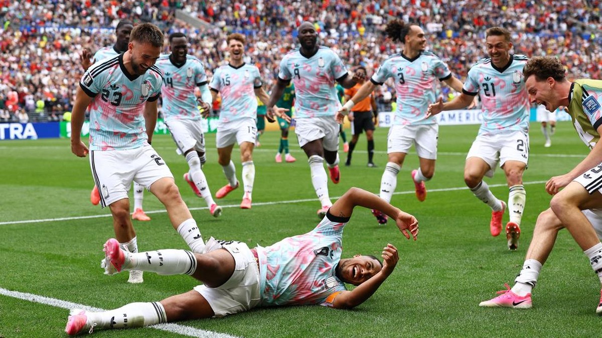 Soccer players celebrate on the field as one player slides on the grass, teammates running toward him with smiles in a packed stadium.