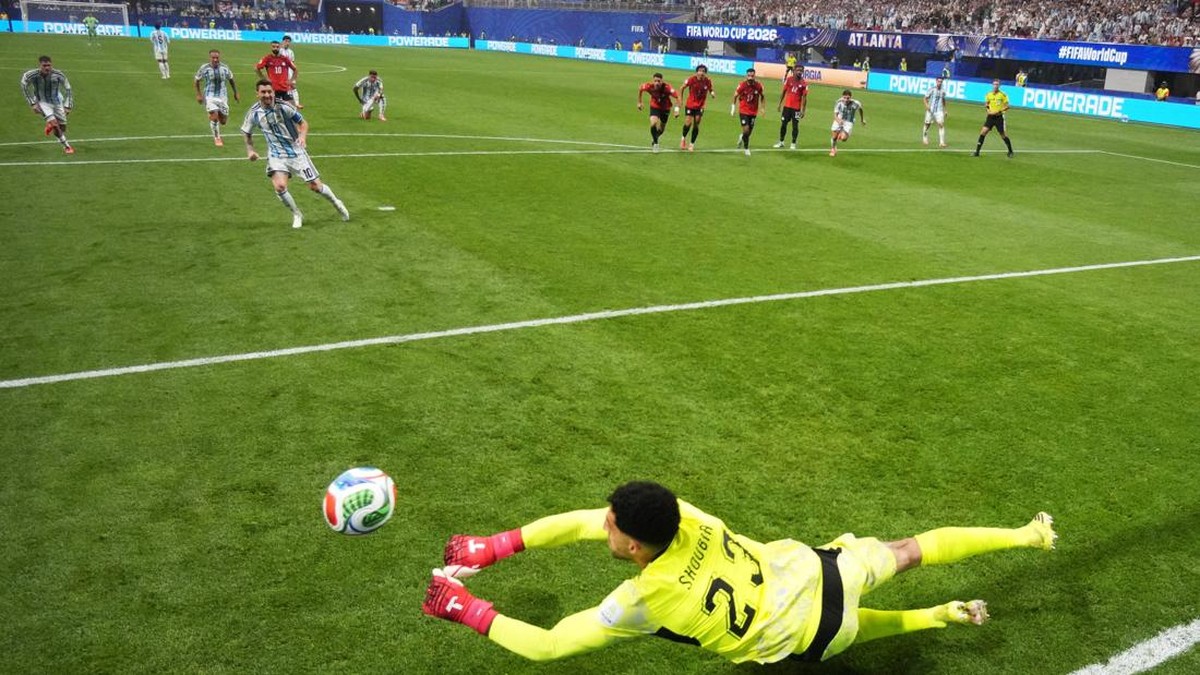 Soccer goalkeeper in yellow dives to save a ball near the bottom left corner as players watch from the penalty area.